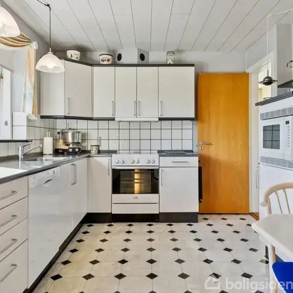 Et hvidt køkken med sorte greb viser en ovn med kogeplader. Skabe og skuffer er langs væggene, og der ses et vindue til venstre. Gulvet er mønstret i sort og hvidt. Der står en hvid stol ved et bord til højre.