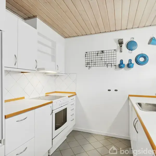 Et hvidt køkken med lysebrunt træloft og flisegulv. Komfuret er i midten med skabe ved siden af. Til højre en vask med vægdekorationer af blå køkkenredskaber. Ingen tekst.