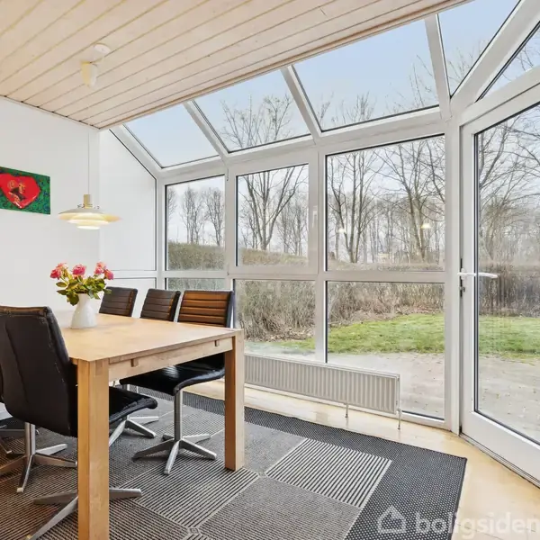 Et spisebord med sorte stole står i en lys og luftig udestue med store vinduer. En vase med blomster pynter på bordet. Udsigt til have og træer.