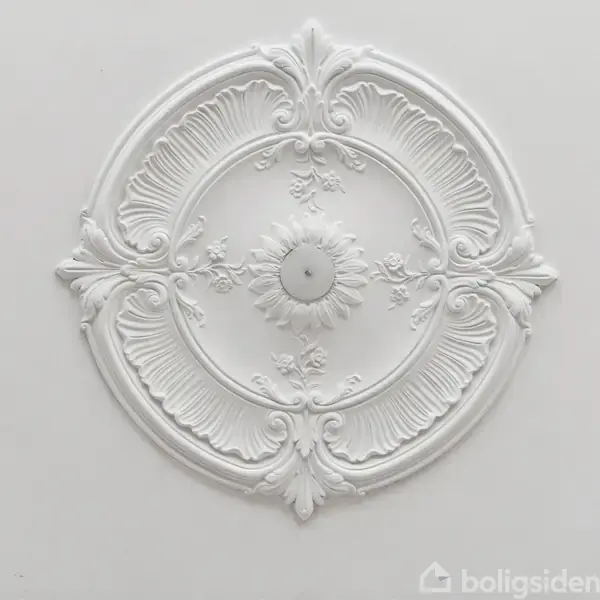 Stukkatur i form af en cirkulær, ornamenteret roset med detaljerede blade og blomster. Den er monteret på en lys loftsflade.