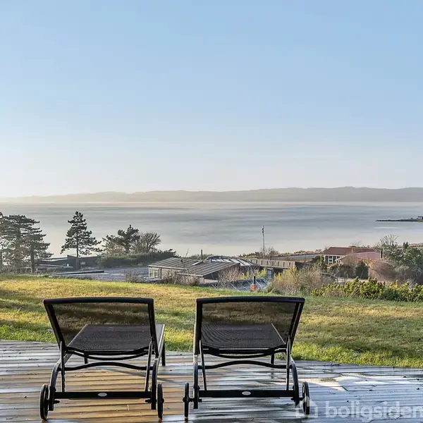 To liggestole står på en træterrasse med udsigt over en rolig kystlinje og grønt landskab. I baggrunden er der hav, træer og bygninger under en klar, blå himmel.