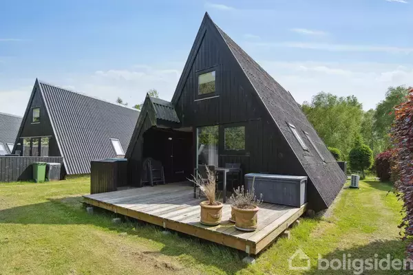 Sort A-formet hus med terrasse, står i solrigt område omgivet af græsplæner og buske. To krukker med planter pryder terrassen. Vinduer på begge etager. Ingen tekst findes i billedet.