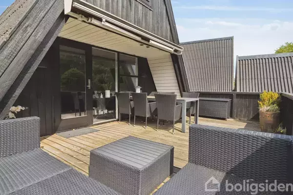 Terrasse med sorte havemøbler står på trædæk. To stole og et bord er placeret ved siden af store glasdøre. Omkringliggende sorte husfacader ses under en blå himmel.