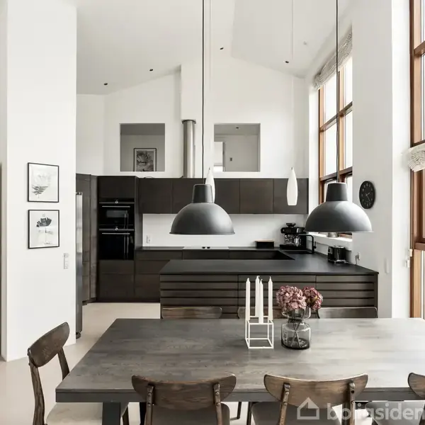 Køkken-alrum med spisebord som hovedobjekt, omgivet af stole. To store lamper hænger over bordet. I baggrunden ses en moderne køkkenindretning med hvidevarer. Store vinduer slipper lys ind.