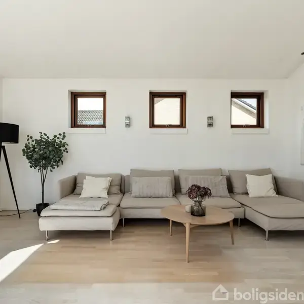 En beige hjørnesofa står i en lys stue med lyse trægulve. Ved siden af er et sort vitrineskab og en stor gulvlampe. Tre små vinduer lyser rummet op, og en abstrakt kunstmaleri hænger på væggen. Et lavt rundt bord står foran sofaen med en vase.