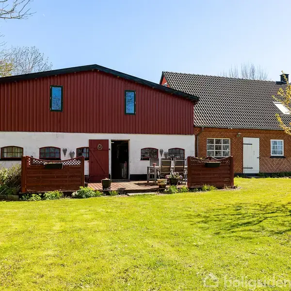 Hus med røde og hvide vægge og sort tag. Foran huset er en terrasse med havemøbler og en velplejet græsplæne. Træer og buske omkranser området, der bades i solskin.