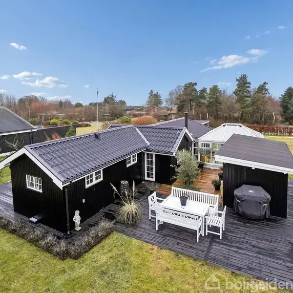 Et hvidt køkkenrum indeholder et moderne komfur med emhætte. En rødorientalsk tæppe pryder gulvet, og hylder med diverse krydderier dekorerer væggen. Vinduer giver naturligt lysindfald.