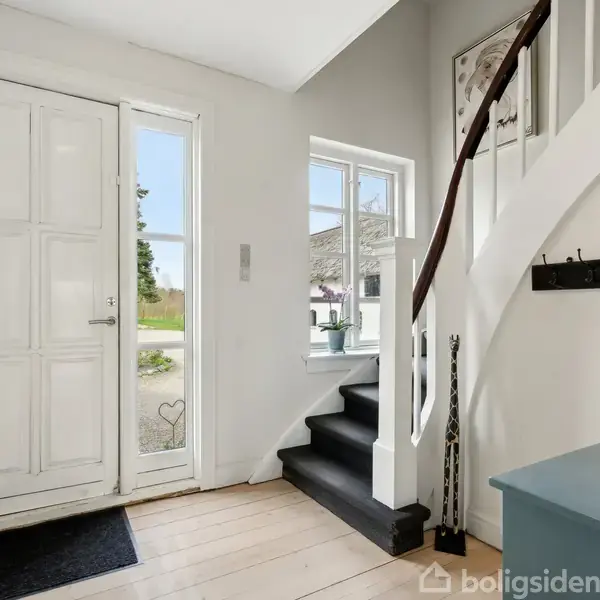 En lys entré med trappe, der fører opad. Yderdøren er hvid med små ruder ved siden af. En blå bænk og knager hænger på væggen. Skilt tekst: "På loftet sidder...".