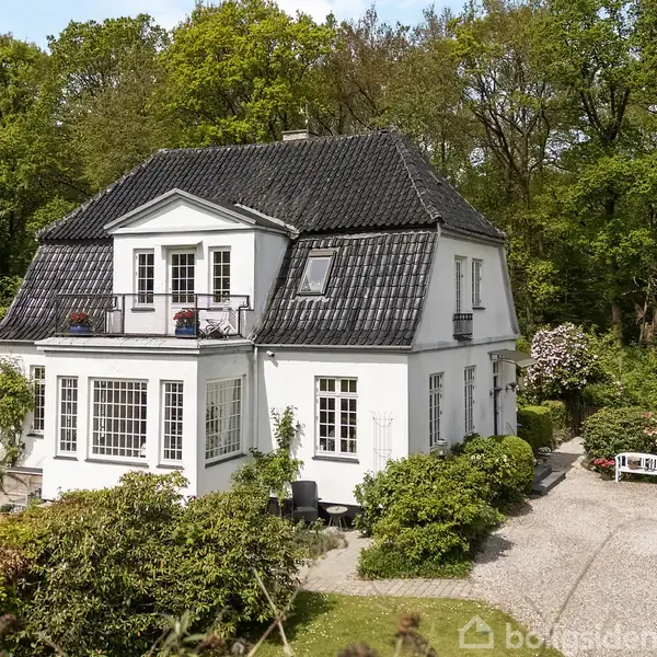 Et hvidt, to-etagers hus med sorte tagsten står omgivet af frodig, grøn have med blomstrende buske. En grussti fører til hoveddøren. Sollys kaster skygger over huset.