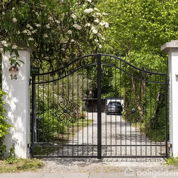 Sort smedejernslåge står åben, fører til en grusvej omgivet af frodig vegetation; husnummer 14 er synligt på venstre søjle. I baggrunden ses en bil.