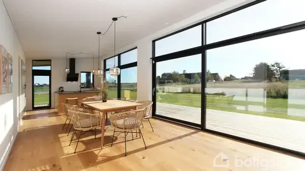 Spisebordet står ved store vinduer, som giver udsigt til haven. I baggrunden ses et moderne køkken. Bordet er omgivet af stole, og rummet er oplyst af naturligt lys.