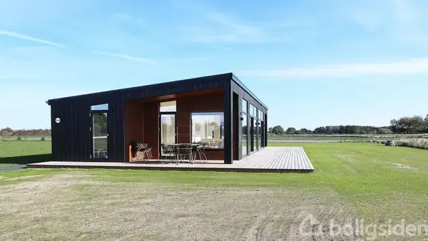 En moderne, sort træhytte står på en grøn mark, med store vinduer og en terrasse med havemøbler. Den åbne himmel skaber en følelse af rummelighed og ro.