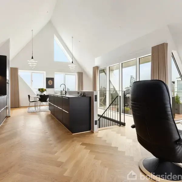 Stue med sort lænestol, køkkenø, og spisebord. Rummet har skråt loft, parketgulv og store glasdøre, der fører til altan. Skandinavisk design med moderne indretning og planter.