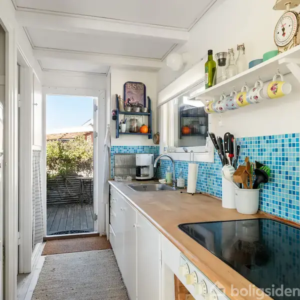 Køkkenbord med induktionskogeplade og opvaskebakke ved et vindue. Farverigt mosaikflise look som backsplash. Hylder med kopper og krydderier. Dør åbner mod terrasse og have.