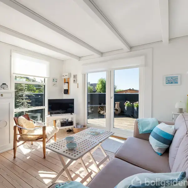 Stue med sofa og sofabord foran dobbeltdøre til terrasse. En stol står ved siden af en hvid reol, og et TV står på et lille bord. Rummet er lyst og elegant indrettet.