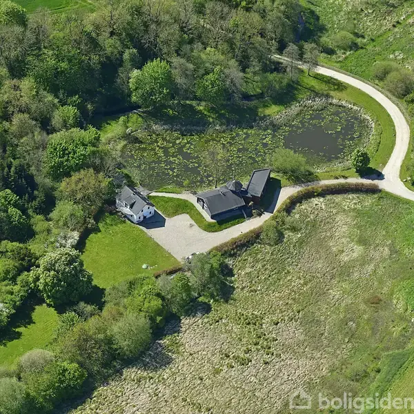 Et hus beliggende ved en sø omgivet af skov og marker. En vej snor sig gennem landskabet, og flere træer omkranser området.