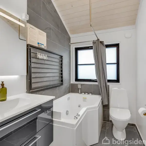 En moderne badeværelsessuite med hvide vægge og gråt flisegulv. Indeholdende et badekar ved siden af et vindue, håndvask med skabsplads og et toilet. En brun flaske står ved vasken.