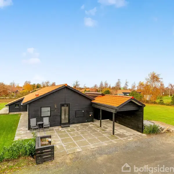 Sort træhus med rødt tegltag står på en flisebelagt terrasse. To stole er placeret foran huset. Græsplæne og træer omkranser huset under en blå himmel.