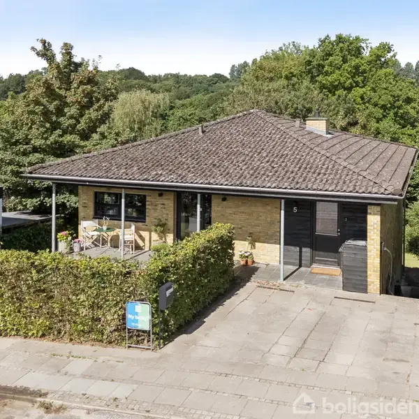 En etplans villa med mørkt tag og gul murstensfacade står omgivet af buske og træer. Foran huset er en betonsti og en skilt. Stille boligområde med natur baggrund.