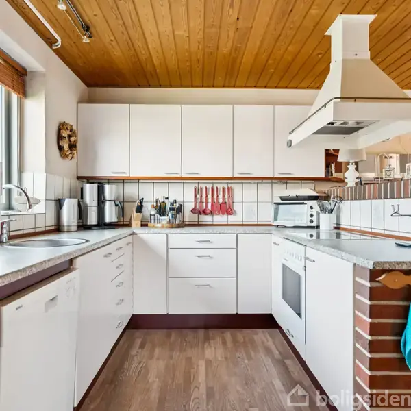 Køkken i L-form med hvide skabe og træloft. Bordplader af grå laminat. Diverse køkkenudstyr placeret på bordet, ovn indbygget i murstenssektion. Vindue lader naturligt lys ind.