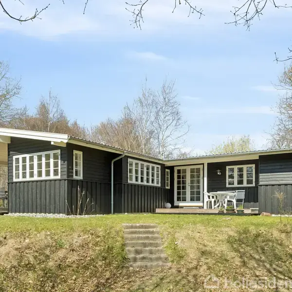 Sort træhus med hvide vindueskarme står stille. Det ligger i et grønt, skovklædt område med græs og træer. Terrasse med havemøbler foran huset.