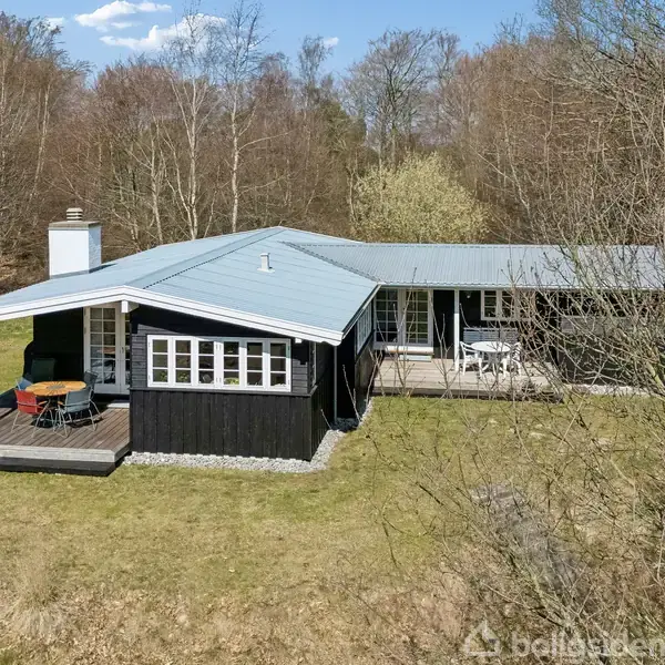 Et sort sommerhus med blåtag og terrasse står i skovklædt område. Der er borde og stole på terrassen. Baggrunden er domineret af træer og græsplæne.