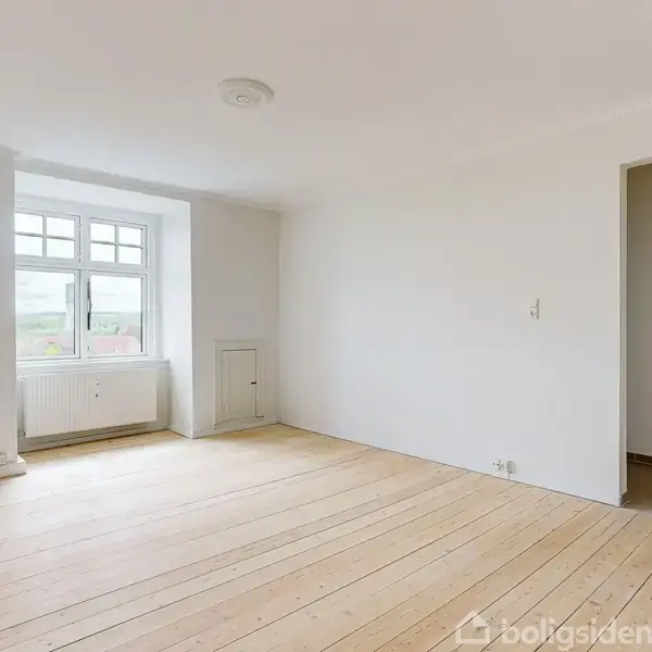 Køkken med grå skabe og stålbordplade, hvor der er en vask. To skråvinduer giver lys. I baggrunden ses en åben dør mod et lyst rum.