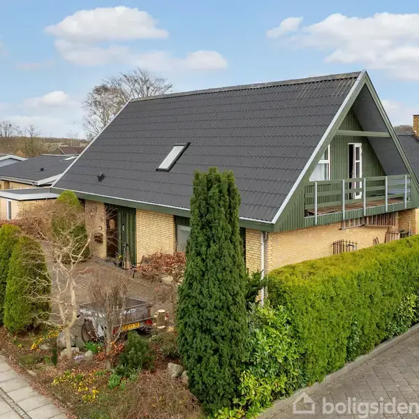 Huset har et A-formet tag med en balkon og ovenlysvindue, omgivet af en velplejet have med træer og buske, beliggende i et parcelhuskvarter med flere huse i baggrunden.