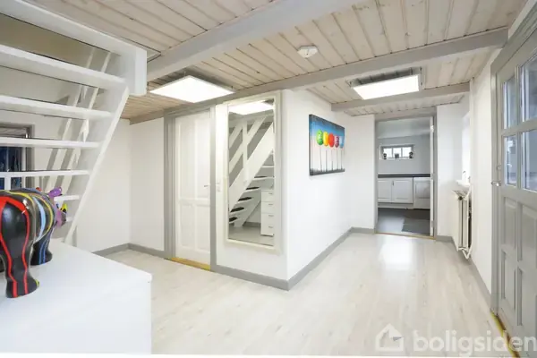 Et lyst rum med trappe til venstre og farverige hoveder på en kommode til venstre. Væggene er prydet med kunst, og en dør leder til et tilstødende køkken.