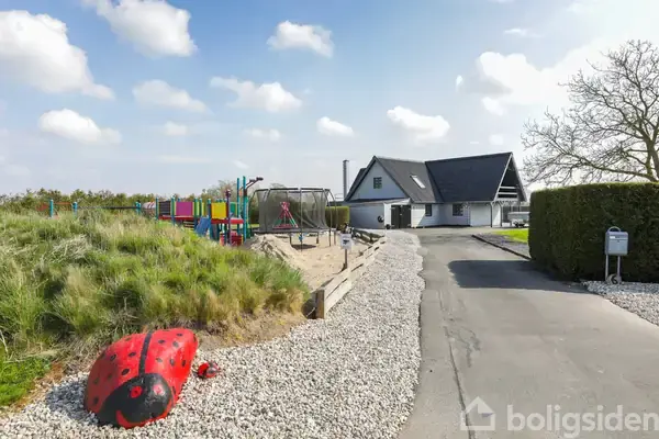 Rød mariehønefigur ligger stille på grus ved siden af en legeplads med gynger og rutsjebane, tæt på et stort hvidt hus. Solrig dag, blå himmel med få skyer.