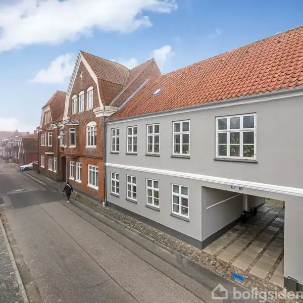 Et gråt hus med rødt tegltag står i en bygade. Ved siden af er røde murstensbygninger. En person går på fortovet ved siden af huset. Bygaden strækker sig ud i det fjerne.