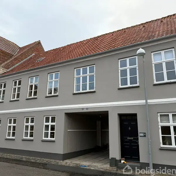 Grå murstensbygning med rødt tegltag, flere hvide vinduer og en sort dør. En åben port forbinder til en gårdsplads. En gadelampe står foran bygningen.