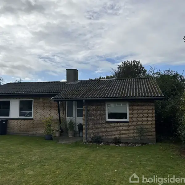 En enkeltstående etplans murstensvilla med skorstensrør på taget står stille. Den er omgivet af en grøn plæne og tæt bevoksning under en overskyet himmel.