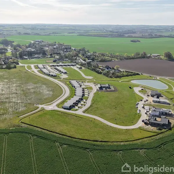Dronebillede af et nyt boligområde under udvikling; veje og huse spredes mellem grønne marker, med landsbyhuse i baggrunden, omgivet af opdyrkede marker og en lille sø.