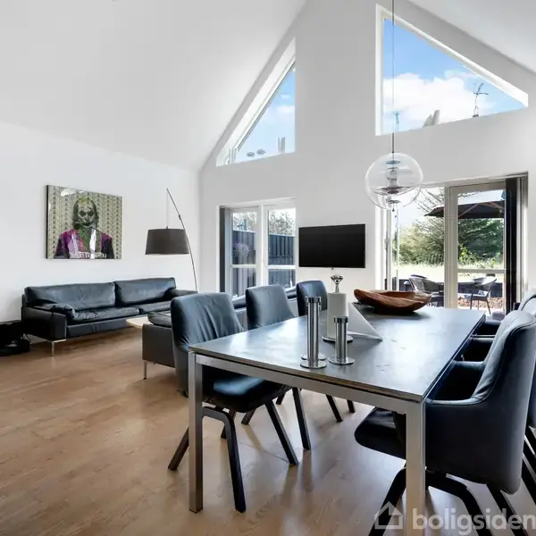 Stuen har et moderne bord med stole, en sofa nær et maleri og en stålampe. Store vinduer og en glaspaneldør leder til en terrasse med udsigt til haven.