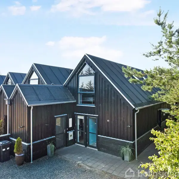 Hus med sort træfacade og blå indgangsdør står stille omgivet af grusvej og grønne træer, mens flere biler er parkeret i nærheden under en blå himmel.