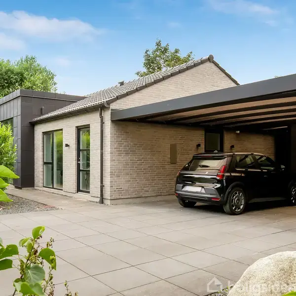 Et moderne hus står i et boligområde, flankeret af grønne buske. En sort bil er parkeret i en åben carport ved siden af huset.