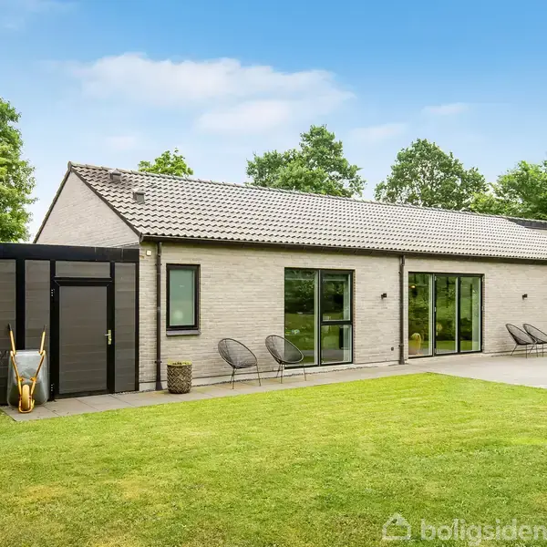 Et moderne hus med fladt tag og store vinduer, omgivet af en velplejet græsplæne og træer. Ved siden af huset står en plæneklipper og en trillebør på en flisebelagt terrasse.