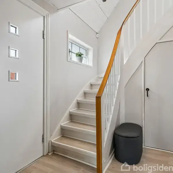 Trappe fører opad, flankeret af gelænder, i en hvidmalet indgang med lyse vægge. En sort skammel står ved siden af trappen. Vindue over trappen lader lys ind.