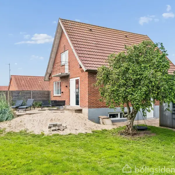 Et rødt murstenshus med skrånende tag står i en have med grøn græsplæne. Omkring huset er der en sandet terrasse med havemøbler og et skur ved siden af.