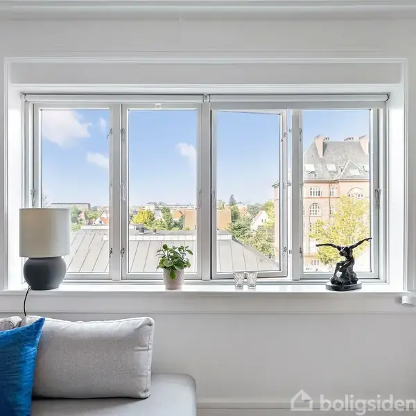 Et vindue med udsigt til bygninger udenfor. På vindueskarmen står en lampe, en grøn plante og en sort skulptur. En grå sofa med en blå pude ses foran.