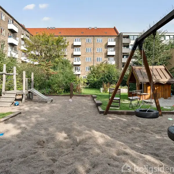 Legeplads med gyngestativ og træhytte i sandet, omgivet af lejlighedsbygninger og grønne træer i en urban gårdhave.