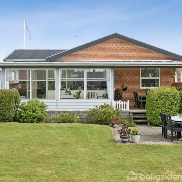 En etplans villa med rød murstensfacade og stort vinduesparti, der omfavner en solrig terrasse. Foran huset er en velplejet have med græsplæne og en lille terrasse med bord og stole.