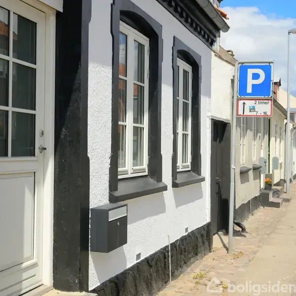 Hvidt og sort byhus med to vinduer, parkering tilladt skilt foran; gade i en række af sammenhængende bygninger under blå himmel. Skilttekst: "2 timer."
