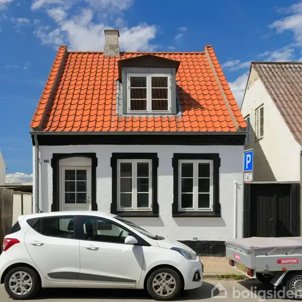 Hus med rødt tegltag og hvid facade med sorte vinduesrammer står ved vej. Hvid bil og trailer parkeret foran. Blå himmel og skilt med "P" i baggrunden.