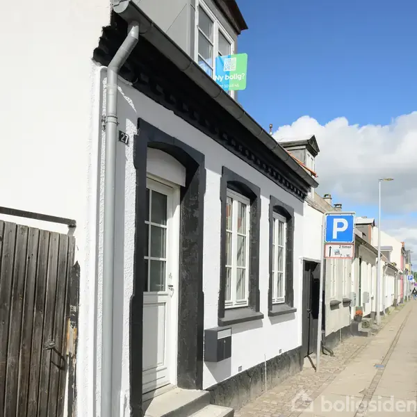 En hvid og sort byhusfacade med murstensdetaljer og tre smalle vinduer. En blå parkeringsskilt står på fortovet ved siden af. Skyer og blå himmel er synlige.