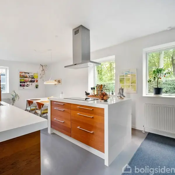 Et køkken med en centralø med træskuffer, en emhætte og hvide bordplader. Rummet har store vinduer med udsigt til en grøn have, og der er moderne belysning.