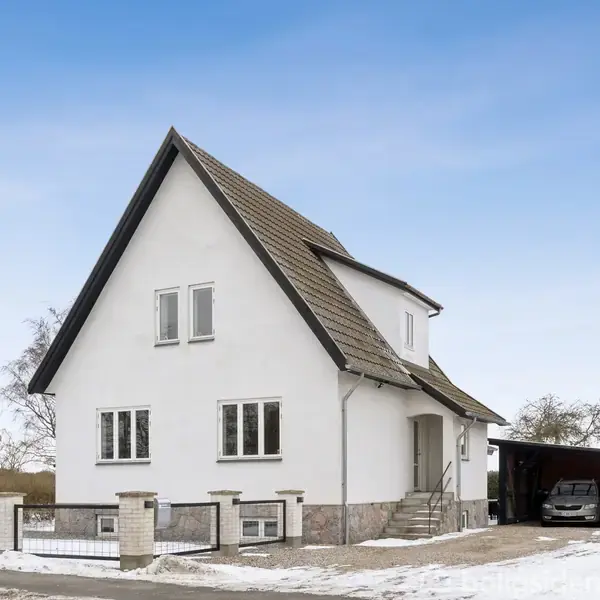 Et hvidt hus med stejlt tag står ved siden af en grusvej. Trappen fører til hoveddøren, og der er en carport med en bil til højre.