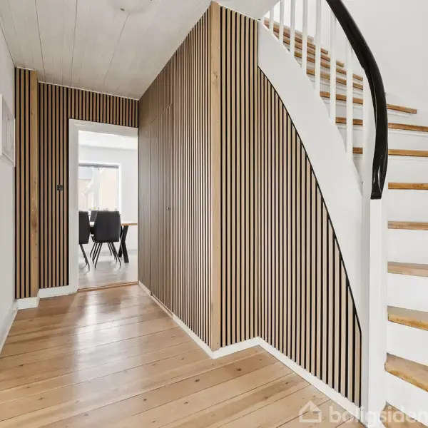 Trappe opad i moderne rum med stribevægge og lyse træplanker, leder til overetage. Gangen åbner op til et rum med sorte stole og store vinduer i baggrunden.