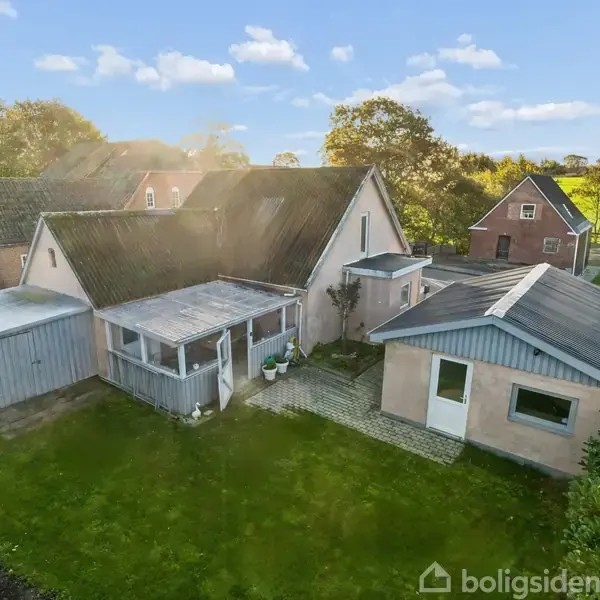 Et parcelhus med flere udhuse er placeret i en grøn have. Et skur er tilknyttet huset. Landskabet er landligt med træer og marker i baggrunden.